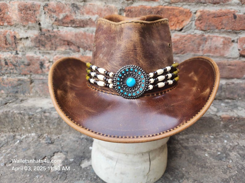 May include: A brown leather cowboy hat with a turquoise and silver concho and a white beaded band.