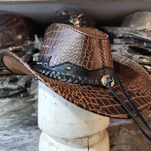 May include: A brown leather cowboy hat with a crocodile pattern and a black braided band. The hat has a silver buckle and a long brown leather strap.