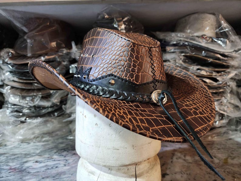 May include: A brown leather cowboy hat with a crocodile pattern and a black braided band. The hat has a silver buckle and a long brown leather strap.