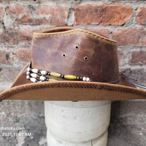 May include: A brown leather cowboy hat with a brown brim and a beaded band. The hat is on a white mannequin head.
