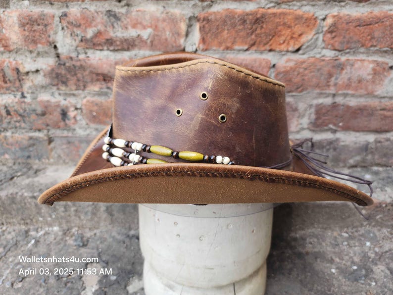 May include: A brown leather cowboy hat with a brown brim and a beaded band. The hat is on a white mannequin head.