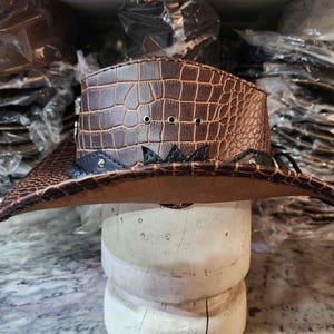May include: A brown leather cowboy hat with a crocodile pattern and black accents. The hat has a wide brim and a tall crown.