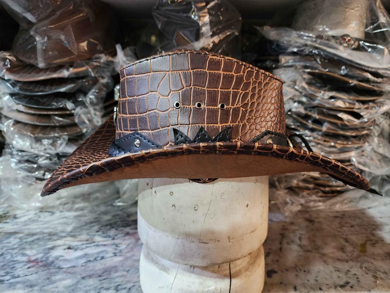 May include: A brown leather cowboy hat with a crocodile pattern and black accents. The hat has a wide brim and a tall crown.