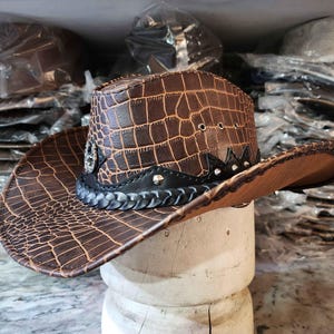 May include: A brown leather cowboy hat with a crocodile pattern and silver accents. The hat has a black braided band and a wide brim.