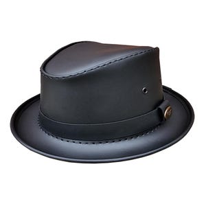 Könnte beinhalten: Ein schwarzer Fedora-Hut aus Leder mit einer abgerundeten Krone und einer kurzen Krempe. Der Hut hat ein dunkles Band um die Basis der Krone und einen dekorativen Knopf an der Seite. Der Hut hat ein klassisches, zeitloses Design.