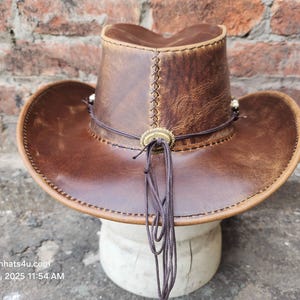 May include: A brown leather cowboy hat with a wide brim and a decorative band. The hat has a distressed look and is made of genuine leather.