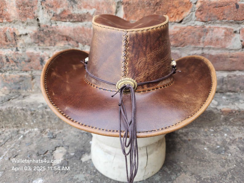 May include: A brown leather cowboy hat with a wide brim and a decorative band. The hat has a distressed look and is made of genuine leather.