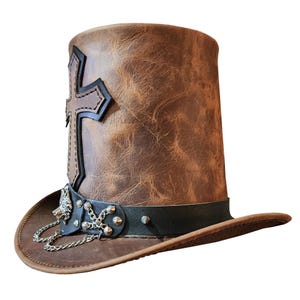 May include: A brown leather top hat with a black band and silver chain accents. The hat features a large, stitched leather cross on the front.