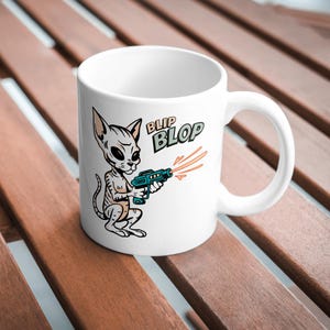 May include: White ceramic mug with a cartoon alien cat character holding a teal ray gun, with the words "BLIP BLOP" in green and black. The alien cat is light grey with large black eyes. Orange laser beams emanate from the gun.