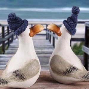 Puede incluir: Dos figuras de cerámica de gaviota con cuerpos blancos, alas grises y picos naranjas. Cada gaviota lleva un gorro de punto azul marino. El fondo muestra una escena de playa con una pasarela de madera y el mar.