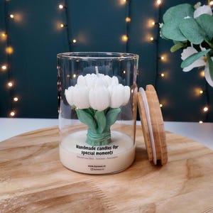 Op de afbeelding: Een kaars in een heldere glazen pot met een witte, tulpvormige wasfiguur met groene stelen. De pot heeft een bamboe deksel en een label met de tekst Handmade candles for special moments, Natural Soy Wax, Scented, Decorator, www.bymasi.nl en @bymasi.nl.