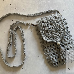 Puede incluir: Una bolsa de teléfono de ganchillo gris con un diseño floral y un borla con cuentas. La bolsa está unida a una larga correa de ganchillo gris.