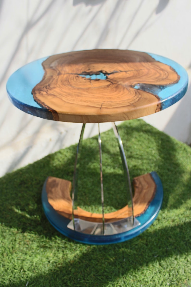 K&ouml;nnte beinhalten: Ein runder Holztisch mit einer blauen Epoxidharzplatte und einem silbernen Metallfu&szlig;. Der Tisch steht auf einer gr&uuml;nen Grasfl&auml;che.