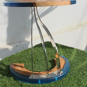 K&ouml;nnte beinhalten: Ein runder Holztisch mit einer blauen Epoxidharzplatte und einem einzigartigen, geschwungenen Edelstahlfu&szlig;. Der Tisch steht auf einer gr&uuml;nen Kunstgrasoberfl&auml;che.
