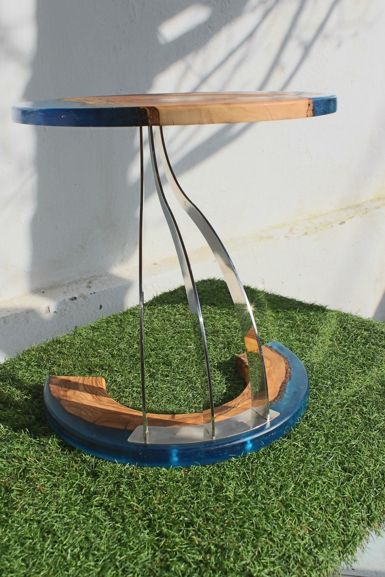 K&ouml;nnte beinhalten: Ein runder Holztisch mit einer blauen Epoxidharzplatte und einem einzigartigen, geschwungenen Edelstahlfu&szlig;. Der Tisch steht auf einer gr&uuml;nen Kunstgrasoberfl&auml;che.