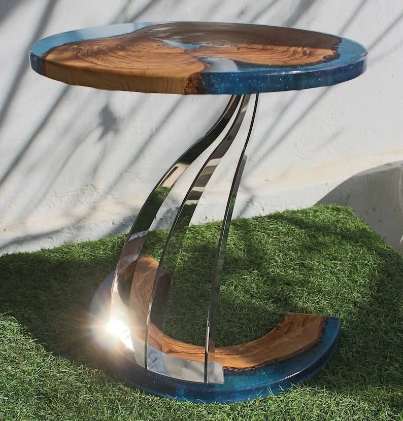 K&ouml;nnte beinhalten: Ein runder Holztisch mit einer blauen Epoxidharzplatte und einem einzigartigen, gedrehten Edelstahlfu&szlig;. Der Tisch steht auf einer gr&uuml;nen Grasfl&auml;che.