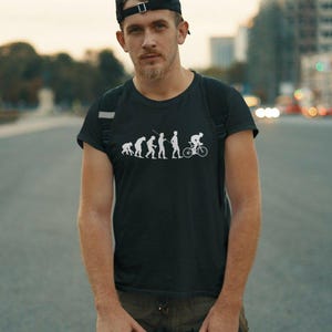 Può includere: Una t-shirt nera a maniche corte con una stampa grafica bianca della sequenza dell'evoluzione umana, che progredisce da un primate a un essere umano in bicicletta. La maglietta ha un girocollo ed è realizzata in tessuto nero.