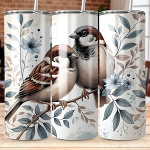 May include: Two sparrows perched on a branch with blue and brown leaves. The sparrows are brown and white with a light blue background. The design is on a white tumbler.