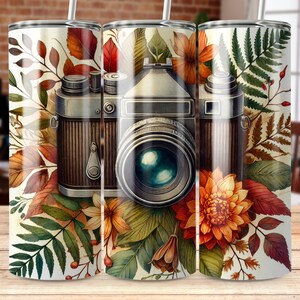 May include: A white tumbler with a vintage camera and autumn leaves design. The camera is brown and has a large lens. The leaves are in shades of red, orange, yellow, and green.