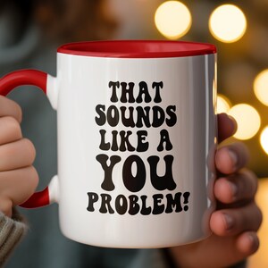 May include: White ceramic mug with a red rim, featuring the text "That sounds like a you problem!" in black lettering. The mug is being held by a person's hand.