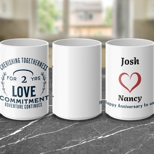 Może przedstawiać: Dwa białe ceramiczne kubki z spersonalizowanym napisem. Pierwszy kubek ma napis "Cherishing Togetherness for 2 yrs Love Commitment Adventure Continues". Drugi kubek ma napis "Josh Nancy Happy Anniversary to us" z czerwonym sercem.
