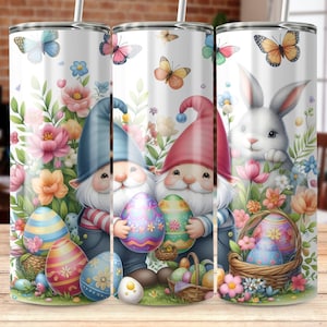 Könnte beinhalten: Drei Edelstahlbecher mit weißem Hintergrund und bunten Ostermotiven. Die Designs zeigen Gnome, Schmetterlinge, Blumen und Ostereier. Ein Becher zeigt ein Kaninchen.