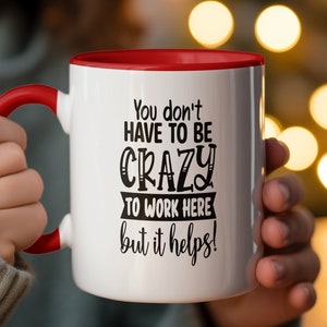 Op de afbeelding: Een witte keramische mok met een rode rand en handvat. De mok heeft zwarte tekst met de tekst "You don't have to be crazy to work here but it helps!"