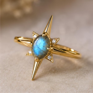 May include: A gold ring featuring a central oval-shaped labradorite gemstone with a blue iridescent sheen. The stone is set within a starburst-shaped frame with four long pointed rays and four small round clear accent stones at the base.