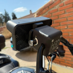 Può includere: Accessorio nero per moto con la scritta "H3D" sul lato. È dotato di una serratura a chiave ed è montato sul manubrio. Lo sfondo comprende un muro di mattoni e una sella per moto.