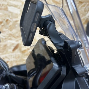 Puede incluir: Un soporte de teléfono para motocicleta negro con un parabrisas de plástico transparente. El soporte está fijado al manillar y tiene un brazo giratorio que se puede ajustar a diferentes ángulos. El teléfono se mantiene en su lugar de forma segura mediante una abrazadera.
