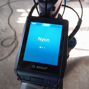 Könnte beinhalten: Ein schwarzes Bosch Nyon E-Bike-Display mit einem blauen Bildschirm, auf dem das Wort "Nyon" und eine Entfernung von 0,0 Meilen angezeigt werden. Das Display ist an einem schwarzen Fahrradlenker montiert.