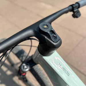 May include: A matte black bicycle handlebar and stem assembly mounted on a light sage green frame. The stem features a circular top cap with a central gold-colored bolt. The frame displays the word Spark in a stylized font near the head tube.