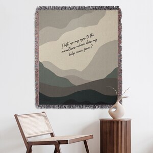 Può includere: Un arazzo tessuto con uno sfondo beige e un design stilizzato di una catena montuosa in tonalità di verde e marrone. L'arazzo presenta il testo "Alzo gli occhi verso le montagne - da dove viene il mio aiuto?"