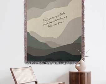 May include: A woven tapestry with a beige background and a stylised mountain range design in shades of green and brown. The tapestry features the text "I lift up my eyes to the mountains - where does my help come from?"