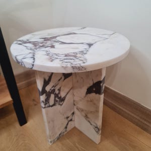 May include: A round white marble side table with a white marble base. The base has a unique design with four legs that are connected in the middle.
