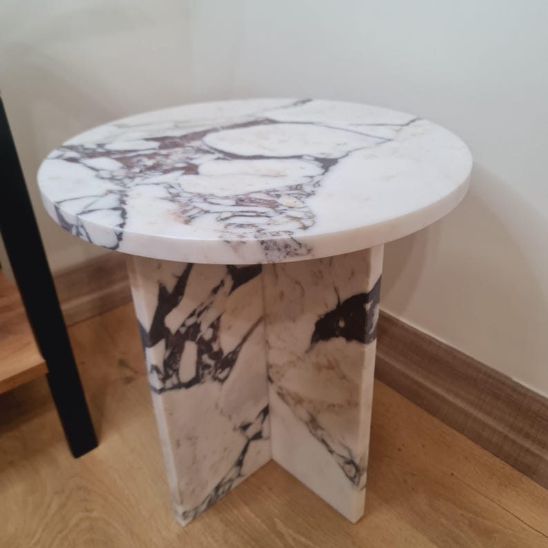 May include: A round white marble side table with a white marble base. The base has a unique design with four legs that are connected in the middle.