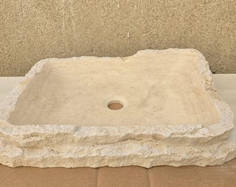 May include: A rectangular, light beige stone sink with a rough, textured exterior. The sink has a central drain hole and a natural, organic shape, suggesting a rustic or natural aesthetic. The sink is made of stone.