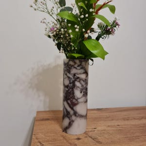 Könnte beinhalten: Eine zylindrische Marmorvase mit einem weißen und violetten Marmormuster. Die Vase ist mit einem Strauß weißer und rosa Blumen und grünen Blättern gefüllt.