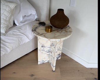 Puede incluir: Una mesa auxiliar de mármol con tapa redonda y base única. La mesa presenta un patrón de mármol blanco y gris. Un jarrón marrón y un pequeño recipiente dorado están encima. La mesa tiene unos 50 cm de diámetro.
