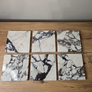 May include: Six square marble coasters with a white background and dark grey and purple veining. The coasters are arranged on a wooden surface, showcasing the natural stone patterns. Each coaster is approximately 10 cm square.