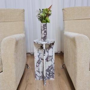 May include: A white marble side table with a round top and a unique, geometric base. The table has a purple and white marbled pattern. A vase with white and purple marbling holds a bouquet of white and green flowers.