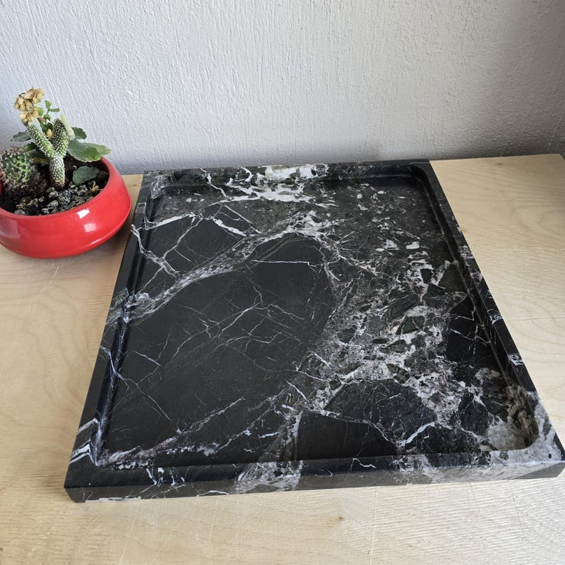 Op de afbeelding: Een rechthoekig dienblad van zwart marmer met opvallende witte aders over het gehele oppervlak. Het dienblad heeft een gladde, gepolijste afwerking met opstaande randen en een platte bodem, wat het natuurlijke steenpatroon mooi weergeeft.