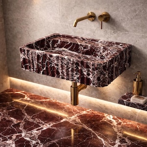 May include: A rectangular, burgundy marble sink with white veining, mounted on a gold-colored stand. A gold faucet and matching fixtures are mounted on the gray tiled wall. A gold soap dispenser and marble soap dish are on the counter.