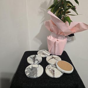 K&ouml;nnte beinhalten: Set aus sechs runden Marmoruntersetzern mit grauer Maserung auf einer schwarzen Oberfl&auml;che. Ein Untersetzer ist umgedreht und zeigt eine Korkbasis. Eine Topfpflanze, eingewickelt in rosa Papier, befindet sich im Hintergrund.