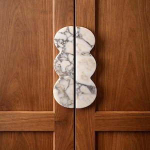 Könnte beinhalten: Nahaufnahme einer Holzschranktür mit einem Paar Marmor-Türgriffen. Die Griffe sind weiß mit grauer Maserung und haben ein einzigartiges, gestapeltes ovales Design. Das Holz hat einen warmen, braunen Farbton.