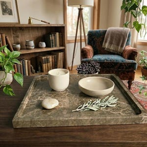 Puede incluir: Una bandeja rectangular con efecto mármol contiene una vela blanca, un cuenco pequeño, una piedra lisa y una ramita de vegetación. La bandeja está sobre una mesa de madera oscura, con un sillón acogedor y una estantería al fondo.