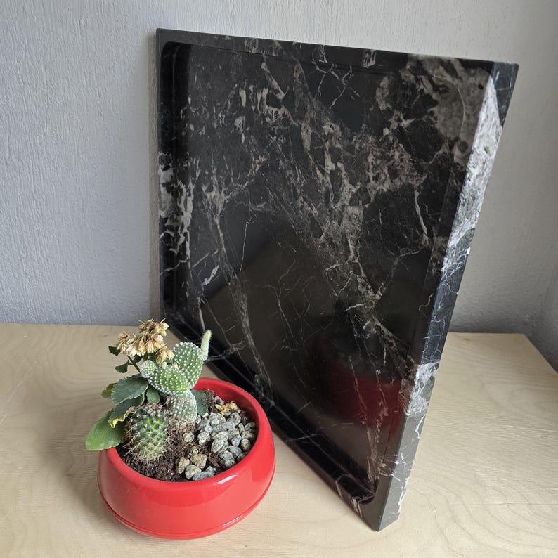 Op de afbeelding: Een vierkant zwart dienblad met een wit marmerachtig aderpatroon en een opstaande rand, geplaatst naast een kleine, ronde, felrode plantenbak met een groene cactus met kleine stekels en wat gedroogde plantenelementen in de aarde.