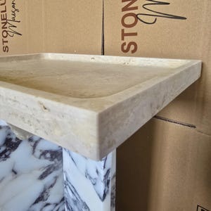 Puede incluir: Una bandeja rectangular de piedra beige con esquinas redondeadas. La bandeja se asienta sobre un pedestal de m&aacute;rmol blanco y gris. El fondo presenta cajas de cart&oacute;n con las palabras "STONELU Maison" y "STON" impresas.