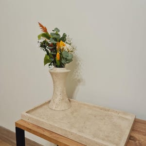 May include: A beige stone tray with a beige stone vase with a bouquet of yellow and white flowers on a wooden table.
