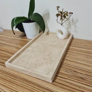 Puede incluir: Una bandeja rectangular de piedra beige con bordes elevados. Un pequeño jarrón blanco con una abertura circular contiene tallos secos. Una orquídea en maceta con hojas verdes está en el fondo. La bandeja está sobre una superficie de madera.